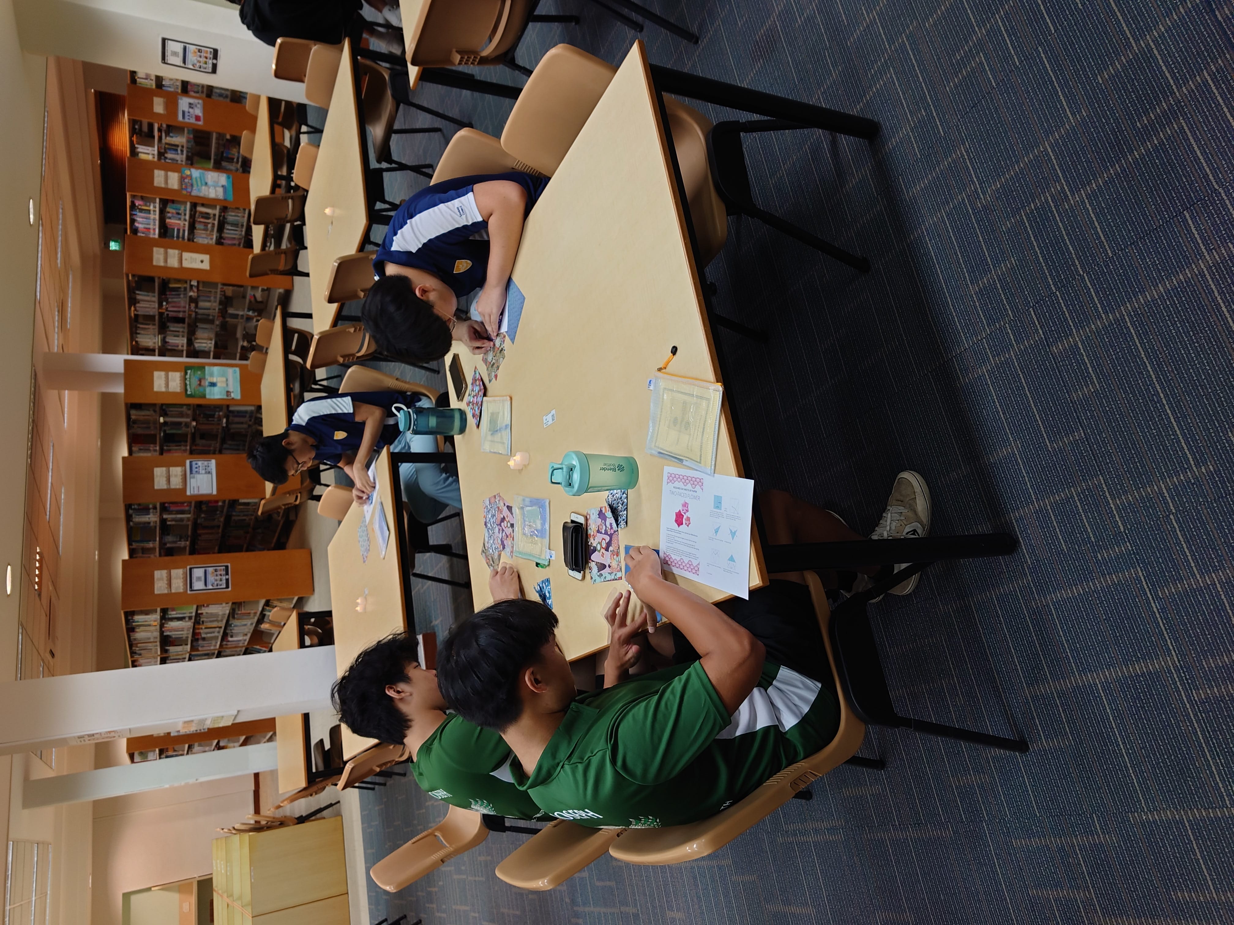 Three students work on papers and cards at a library table, with bookshelves and more tables visible behind them.